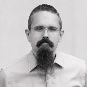 A black and white headshot of a man with glasses, slicked-back dark hair, and a dark mustache and beard.