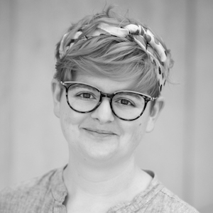 A black and white headshot of a person with rounded glasses and very short hair tied with a scarf, who is looking directly at the camera and smiling slightly.