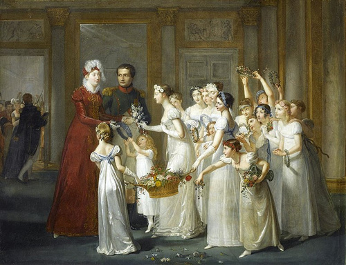 A large group of young women and girls, all wearing white dresses and carrying flowers or wearing flowers in their hair, greet a tall woman in a long-sleeved red dress with a feathered headpiece. Next to the woman in red is a man wearing a blue and red jacket.