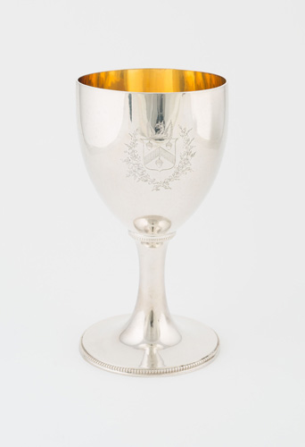 A bright silver goblet with silver beading design around the edge of the foot. The interior is a bright yellow-gold color. The side of the goblet is etched with a coat of arms that includes three acorns, a dragon, and a laurel wreath.