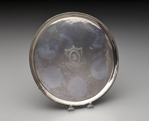 A round, slightly tarnished silver plate or flat dish with a silver beading decoration around teh edge. A coat of arms is etched in the center of the dish. In the tarnish, six circles are visible where something rested on the dish.