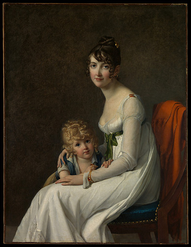 A young woman with light skin and dark curly hair is seated in a side profile, leaning slightly forward. She holds a young child with blonde curls against her with her arm around the child's shoulders. Both lok directly out at the viewer.