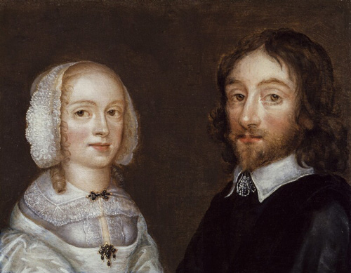 A woman with light skin and blonde curls and a man with light skin, dark hair, and a beard are shown from the shoulders up. They are turned slightly toward each other, but look outward to the viewer.