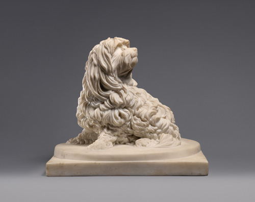 An off-white sculpture of a dog seated on its hind legs, looking back over its shoulder. It has a short snout and long, slightly curled hair all over its body, with longer hair around the head and ears.