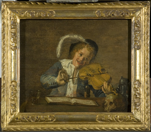 A young man or child with light skin, blonde hair, a feathered cap, and a blue coat is seated at a table and smiling while he plays a violin. On teh table before him is a book that appears to contain sheet music, an empty glass, and a human skull.