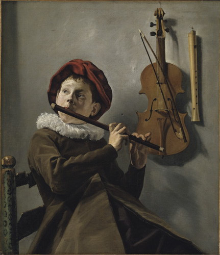 A young boy with very light skin in a brown coat with a large white ruffled collar and red hat is seated in a chair playing a dark-colored flute. Behind him on teh wall hangs a violin and another musical instrument that resembles a white recorder.
