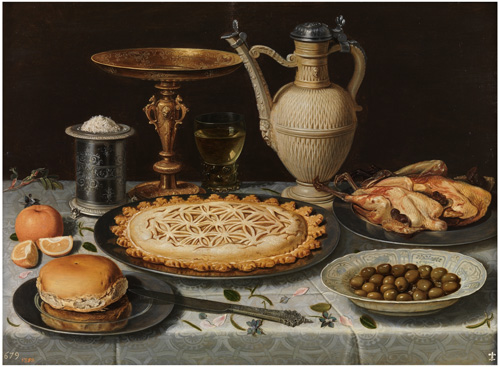 A table with a silvery-blue tablecloth is set with a roast bird, green olives, a pie with a geometric pattern on the top, a bread roll, and an orange. Behind the food, a silver cylinder with salt on top is next to a gold-colored drinking vessel with a very wide, very shallow bowl. On the other side, a wine glass with liquid and a cream-colored pitcher.