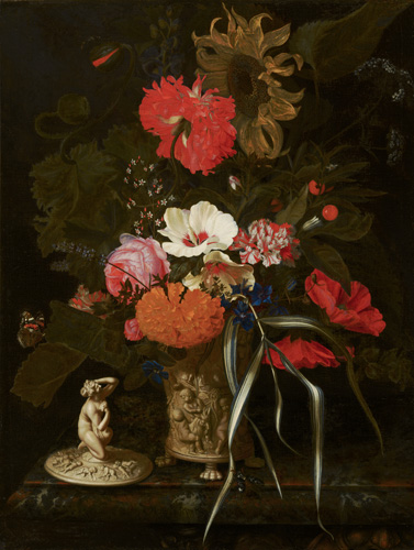 A large flower arrangement sits on a table in a carved off-white vase with a lid resting nearby. The flowers are mostly red and orange, with one large sunflower and a few white flowers at teh center, and a lot of greenery at the back of the vase. The vase is carved with human figures, and the lid has a kneeling nude woman on the top.